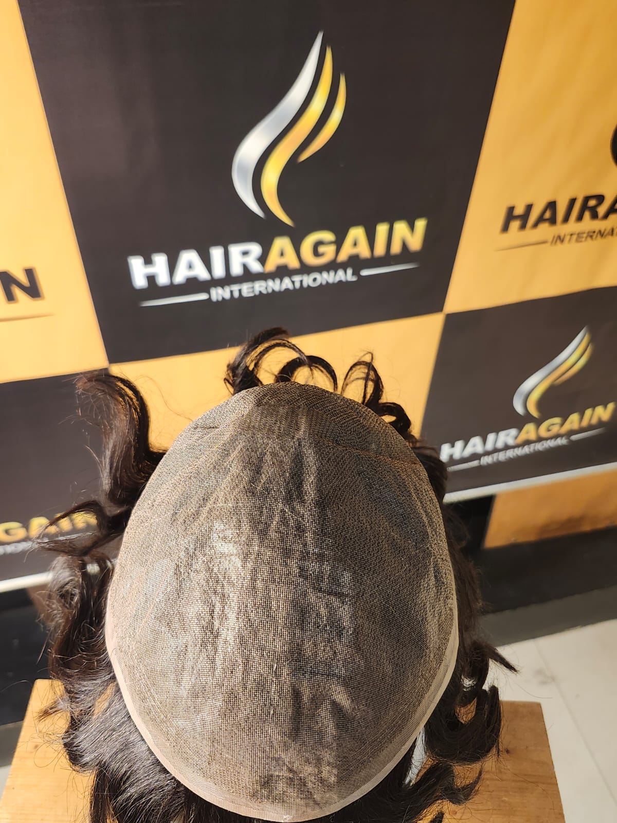 Hairpiece on a wooden block with HairAgain International branding in the background.